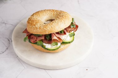 Pastırmalı simit sandviç, salatalık dilimleri, su teresi salatası ve mermer zemin üzerinde ricotta.