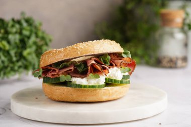 Pastırmalı simit sandviç, salatalık dilimleri, su teresi salatası ve mermer zemin üzerinde ricotta.