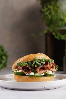 Pastırmalı simit sandviç, salatalık dilimleri, su teresi salatası ve mermer zemin üzerinde ricotta.