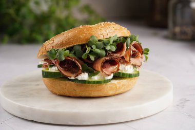 Pastırmalı simit sandviç, salatalık dilimleri, su teresi salatası ve mermer zemin üzerinde ricotta.