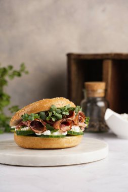 Pastırmalı simit sandviç, salatalık dilimleri, su teresi salatası ve mermer zemin üzerinde ricotta.