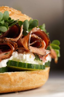 Pastırmalı simit sandviç, salatalık dilimleri, su teresi salatası ve mermer arka planda ricotta, yakın plan, seçici odak