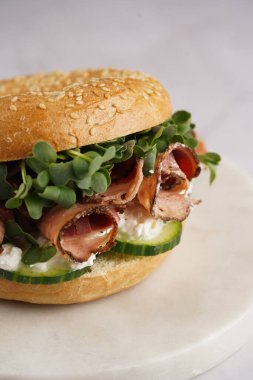 Pastırmalı simit sandviç, salatalık dilimleri, su teresi salatası ve mermer tahtada ricotta.