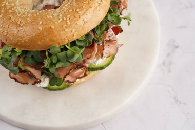 Pastırmalı simit sandviç, salatalık dilimleri, su teresi salatası ve mermer arka planda ricotta.