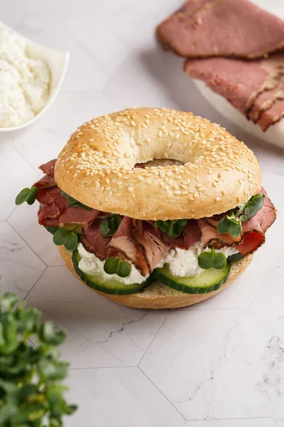 Pastırmalı simit sandviç, salatalık dilimleri, su teresi salatası ve mermer zemin üzerinde ricotta.