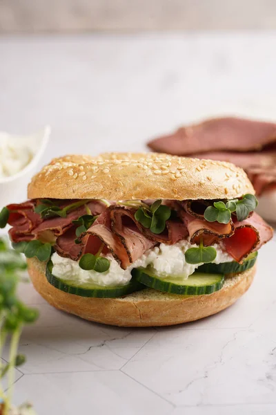 Pastırmalı simit sandviç, salatalık dilimleri, su teresi salatası ve mermer zemin üzerinde ricotta.