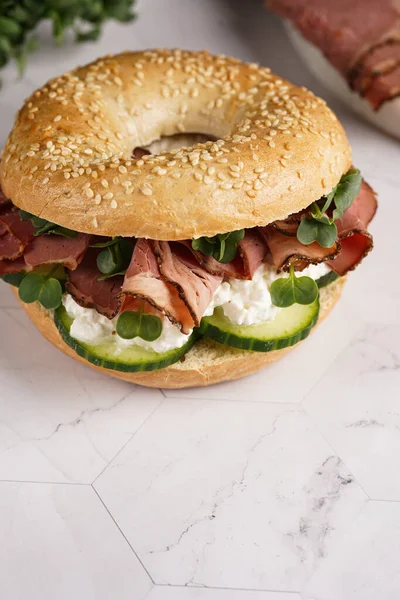 Pastırmalı simit sandviç, salatalık dilimleri, su teresi salatası ve mermer zemin üzerinde ricotta.