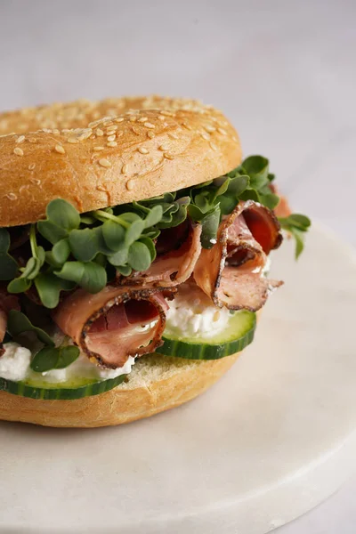 Pastırmalı simit sandviç, salatalık dilimleri, su teresi salatası ve mermer tahtada ricotta.