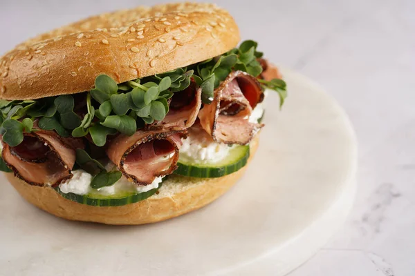 Pastırmalı simit sandviç, salatalık dilimleri, su teresi salatası ve mermer tahtada ricotta.