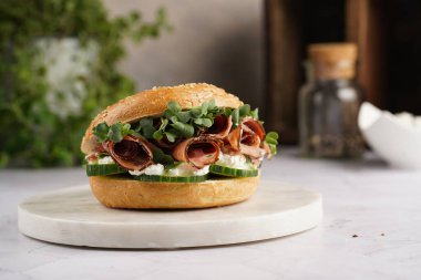 Pastırmalı simit sandviç, salatalık dilimleri, su teresi salatası ve mermer tahtada ricotta.