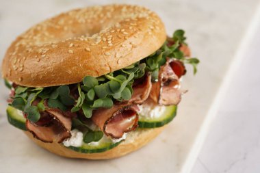 Pastırmalı simit sandviç, salatalık dilimleri, su teresi salatası ve mermer tahtada ricotta.