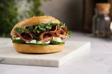 Pastırmalı simit sandviç, salatalık dilimleri, su teresi salatası ve mermer tahtada ricotta.