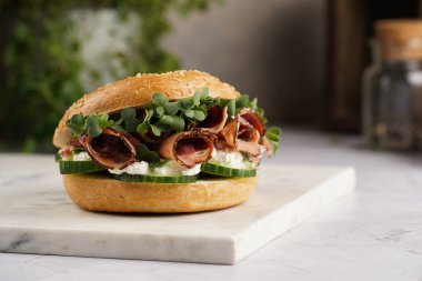 Pastırmalı simit sandviç, salatalık dilimleri, su teresi salatası ve mermer tahtada ricotta.