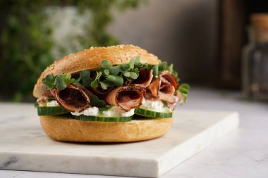 Pastırmalı simit sandviç, salatalık dilimleri, su teresi salatası ve mermer tahtada ricotta.