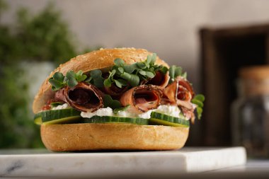 Pastırmalı simit sandviç, salatalık dilimleri, su teresi salatası ve mermer tahtada ricotta.