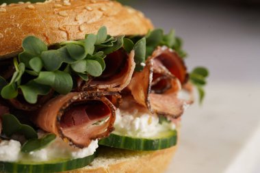 Pastırmalı simit sandviç, salatalık dilimleri, su teresi salatası ve mermer tahtada ricotta, yakın plan, seçici odaklanma.