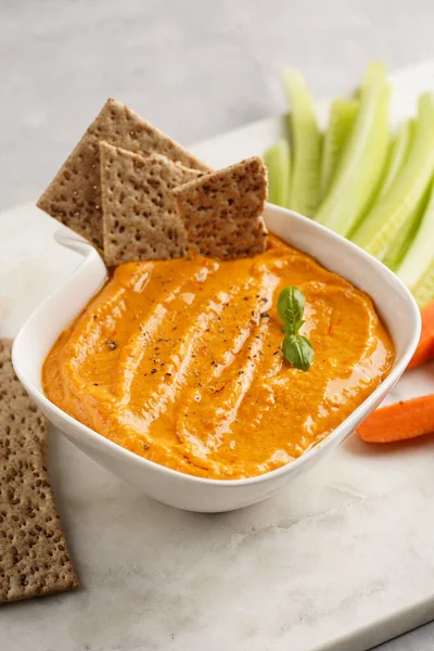 Paprika bell-pepper hummus dip looking like orange pumpkin puree, thin crackers with sesame seeds and carrot and cucumber cut in long strips and sticks on marble board