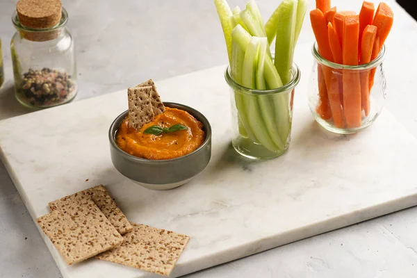 Paprika bell-pepper hummus dip looking like orange pumpkin puree, thin crackers with sesame seeds and carrot and cucumber cut in long strips and sticks on marble board