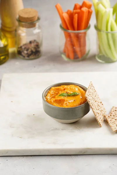 Paprika bell-pepper hummus dip looking like orange pumpkin puree, thin crackers with sesame seeds and carrot and cucumber cut in long strips and sticks on marble board