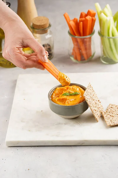 Female hand reaching for paprika bell-pepper hummus dip looking like orange pumpkin puree, thin crackers with sesame seeds and carrot and cucumber cut in long strips and sticks on marble board