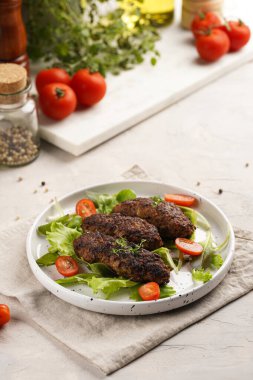 Birkaç ev yapımı kızarmış kıymalı sosis oval bej bir tabakta yeşil yapraklı ve yeşil kareli mutfak havlusunda kiraz domatesli koefte.