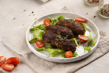 Birkaç ev yapımı kızarmış kıymalı sosis oval bej bir tabakta yeşil yapraklı ve yeşil kareli mutfak havlusunda kiraz domatesli koefte.