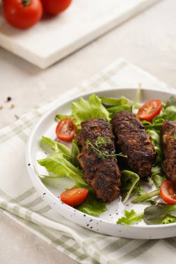 Birkaç ev yapımı kızarmış kıymalı sosis oval bej bir tabakta yeşil yapraklı ve yeşil kareli mutfak havlusunda kiraz domatesli koefte.