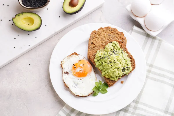 Slices of gluten-free sunflower seeds bread with mashed avocado, fried egg and sesame seeds on white plate on green checkered napkin