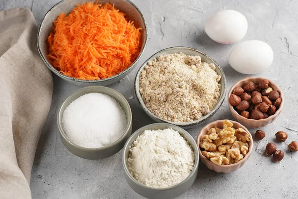 Ingredients for baking a carrot cake: finely grated carrots, grounded hazelnuts and almonds, chopped walnuts, white flour, eggs, sugar on grey background