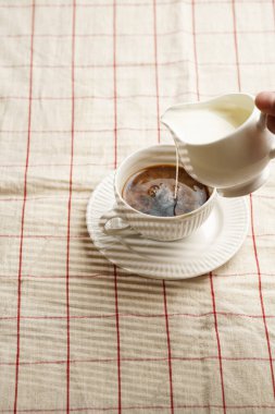 Sütü beyaz porselen bardağa dolduruyor ve kareli masa örtüsüne koyu kahve döküyor.