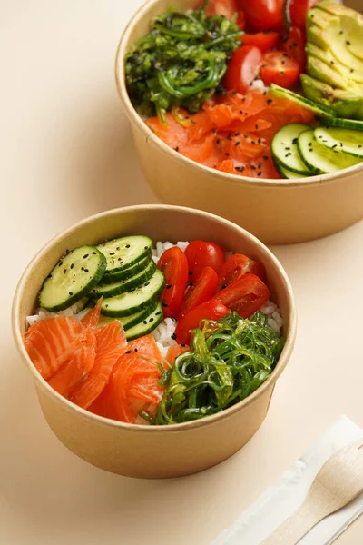 Trendy wholesome dish poke or buddha bowl - rice, wakame seaweed, tomatoes, cucumber, and red fish salmon - in a recycled round carton with sustainable fork, takeaway food concept on beige background