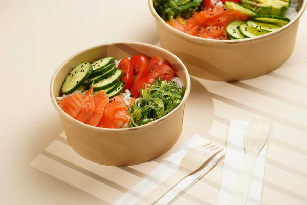 Trendy wholesome dish poke or buddha bowl - rice, wakame seaweed, tomatoes, cucumber, and red fish salmon - in a recycled round carton with sustainable fork, takeaway food concept on beige background