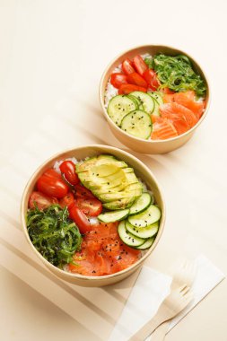 Lezzetli bir tabak dürtme ya da Buda kasesi - pirinç, wakame yosunu, domates, salatalık ve kırmızı balık somonu - sürdürülebilir çatalla geri dönüşümlü yuvarlak bir kutuda, bej arka planda yiyecek kavramı