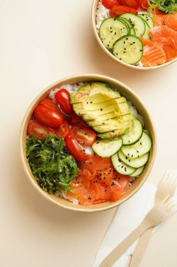 Lezzetli bir tabak dürtme ya da Buda kasesi - pirinç, wakame yosunu, domates, salatalık ve kırmızı balık somonu - sürdürülebilir çatalla geri dönüşümlü yuvarlak bir kutuda, bej arka planda yiyecek kavramı