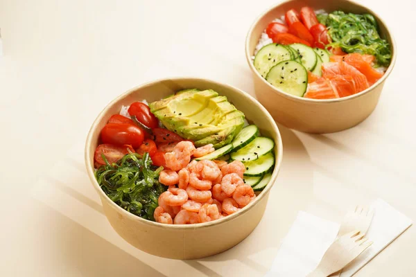 Trendy wholesome dish poke or buddha bowl - rice, wakame seaweed, tomatoes, cucumber, red fish salmon, shrimps - in a recycled round carton with sustainable fork, takeaway food concept