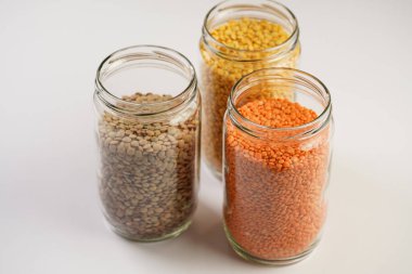 Three tall glass jars with dry red, yellow and green lentils on white background, top view