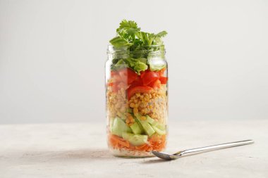 Trendy Fresh detox summer salad layered in a glass mason jar: yoghurt dressing, carrots, cucumber, yellow lentils, red bell pepper and green leaves and herbs, long silver spoon, on white background