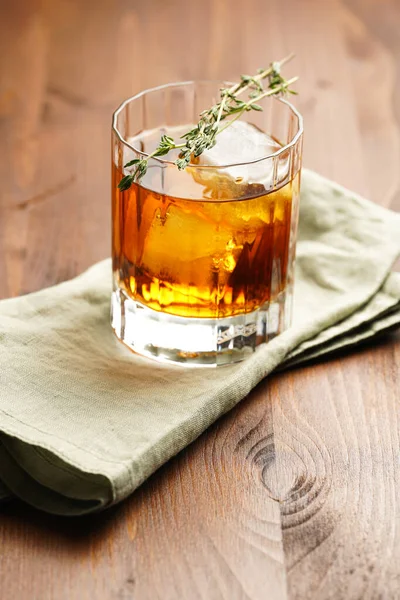 a tumbler glass with brown alcoholic drink, thyme and ice cubes on linen napkin - whisky, rum or cognac - on wooden table, rural scene