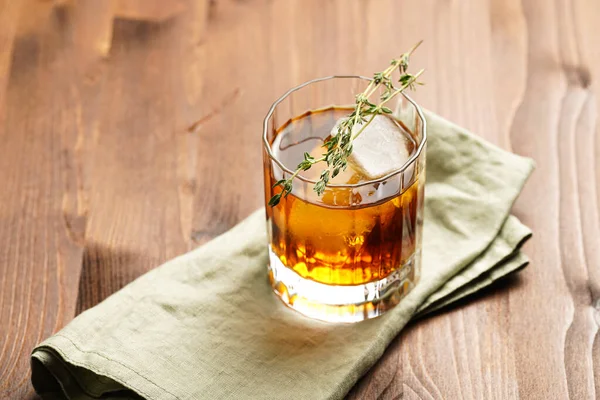 a tumbler glass with brown alcoholic drink, thyme and ice cubes on linen napkin - whisky, rum or cognac - on wooden table, rural scene