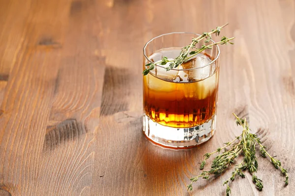 a tumbler glass with brown alcoholic drink, thyme and ice cubes - whisky, rum or cognac - on wooden table, rural scene