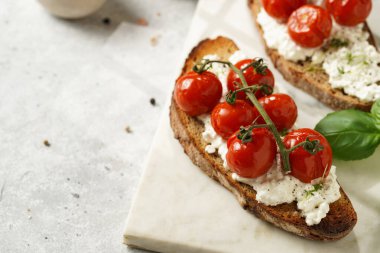 Kızarmış kiraz domatesli sandviç, taze süzme peynir, yeşil fesleğen, gri arka planda mermer bir tahtada kepekli tam buğday ekmeği