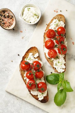 Kızarmış kiraz domatesli sandviç, taze süzme peynir, yeşil fesleğen, gri arka planda mermer bir tahtada kepekli tam buğday ekmeği
