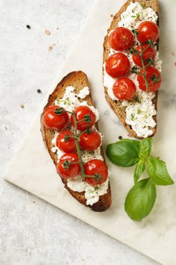 A sandwich with roasted cherry tomatoes with branch, fresh cottage cheese, green basil on a slice of whole wheat bread on a marble board on grey background