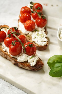 Kızarmış kiraz domatesli sandviç, taze süzme peynir, yeşil fesleğen, gri arka planda mermer bir tahtada kepekli tam buğday ekmeği