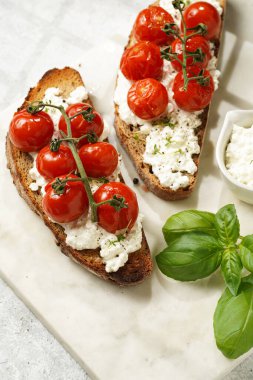 Kızarmış kiraz domatesli sandviç, taze süzme peynir, yeşil fesleğen, gri arka planda mermer bir tahtada kepekli tam buğday ekmeği