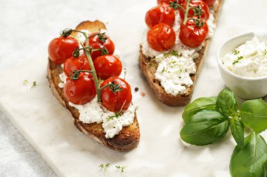 Kızarmış kiraz domatesli sandviç, taze süzme peynir, yeşil fesleğen, gri arka planda mermer bir tahtada kepekli tam buğday ekmeği