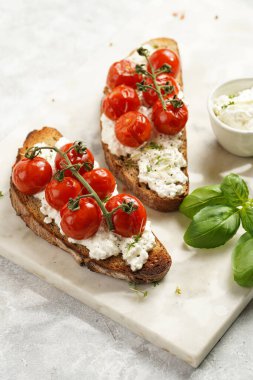 A sandwich with roasted cherry tomatoes with branch, fresh cottage cheese, green basil on a slice of whole wheat bread on a marble board on grey background