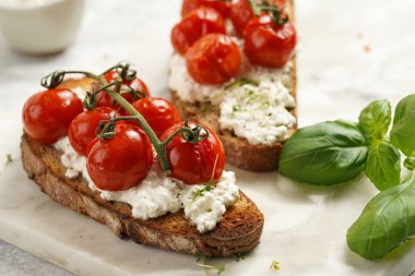 Kızarmış kiraz domatesli sandviç, taze süzme peynir, yeşil fesleğen, gri arka planda mermer bir tahtada kepekli tam buğday ekmeği