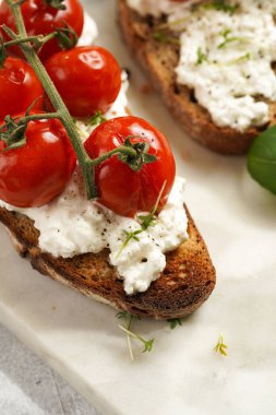Kızarmış kiraz domatesli sandviç, taze süzme peynir, yeşil fesleğen, gri arka planda mermer bir tahtada kepekli tam buğday ekmeği