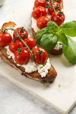 A sandwich with roasted cherry tomatoes with branch, fresh cottage cheese, green basil on a slice of whole wheat bread on a marble board on grey background